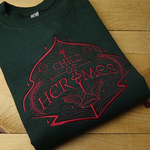 May include: Dark green sweatshirt with a red embroidered design. The design features the text "CHILD OF HERMES" in a decorative font, along with a sword and other ornate elements. The sweatshirt is folded on a wooden surface.