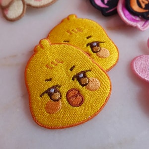 Può includere: Due toppe ricamate gialle con la faccia di un pulcino dei cartoni animati. Il pulcino ha occhi marroni, becco arancione e guance arancioni. Le toppe sono bordate di arancione e hanno un aspetto strutturato.