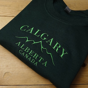 Pode incluir: Um moletom verde escuro com a palavra "CALGARY" arqueada acima de "ALBERTA" e "CANADA" em bordado verde. O design inclui um gráfico de cordilheira. O moletom está dobrado sobre uma superfície de madeira.
