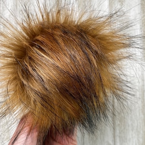 May include: A fluffy pom pom with long, brown and black fur strands. The pom pom is approximately 12.5 cm in diameter.