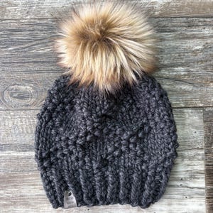 May include: A dark grey knitted beanie with a fluffy brown pom pom on top. The beanie has a woven label attached to the side.