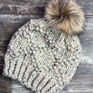 May include: A cream and grey speckled knitted beanie hat with a light brown faux fur pom-pom. The hat has a textured stitch pattern. A warm and stylish winter accessory, perfect for cold weather.