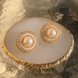 Peut inclure: Une paire de boucles d'oreilles rondes perlées avec une grosse perle centrale. Les boucles d'oreilles ont un cadre doré avec des cristaux irisés et de petites perles blanches. Elles sont présentées sur un plateau décoratif blanc et doré.