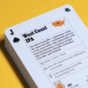 Op de afbeelding: Een stapel witte kaarten met zwarte tekst en illustraties, met informatie over West Coast IPA-bier. De kaarten liggen op een gele ondergrond. De bovenste kaart toont de schoppenboer.