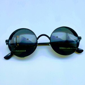 May include: Black round sunglasses with a black frame. The lenses are dark and reflect a blurry image of a building.