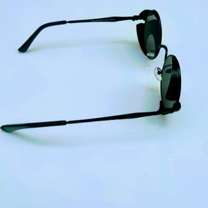 May include: Black round sunglasses with a thin metal frame. The sunglasses have a dark lens and a small metal detail on the side.