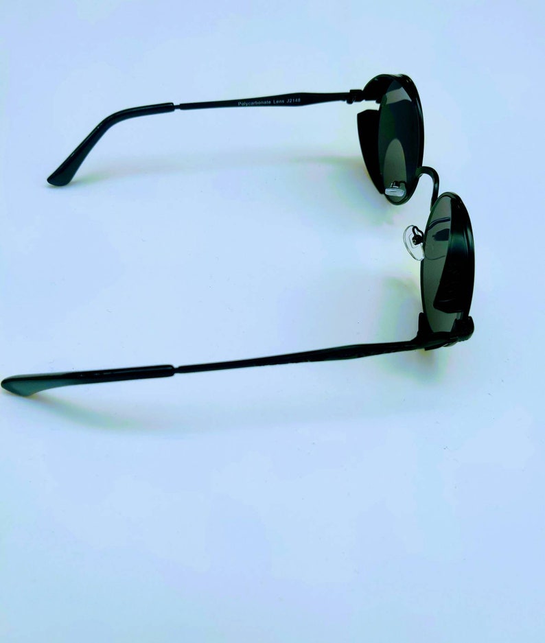 May include: Black round sunglasses with a thin metal frame. The sunglasses have a dark lens and a small metal detail on the side.