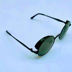 May include: Black round sunglasses with a metal frame and dark lenses. The sunglasses have a unique design with a hinge on the side of the frame.