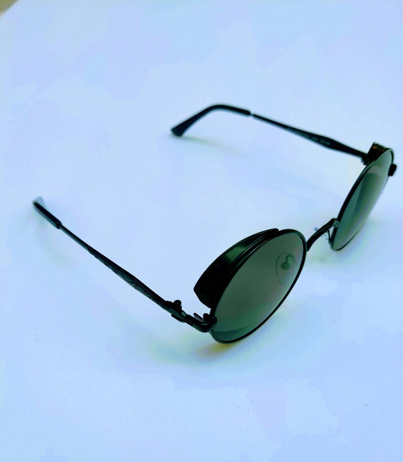 May include: Black round sunglasses with a metal frame and dark lenses. The sunglasses have a unique design with a hinge on the side of the frame.