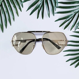 May include: Gold-toned sunglasses with a geometric design. The lenses are a light brown color, and the frames have a double bridge. The sunglasses are set against a white background with green palm leaves.