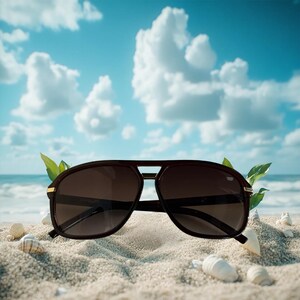 May include: Dark brown aviator sunglasses with a double bridge and tinted lenses rest on a sandy beach. The sunglasses have gold accents and are surrounded by seashells and green leaves. The background features a blue sky with white clouds and the ocean.