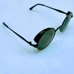 May include: Black round sunglasses with a metal frame and dark lenses. The sunglasses have a unique design with a hinge on the side of the frame.