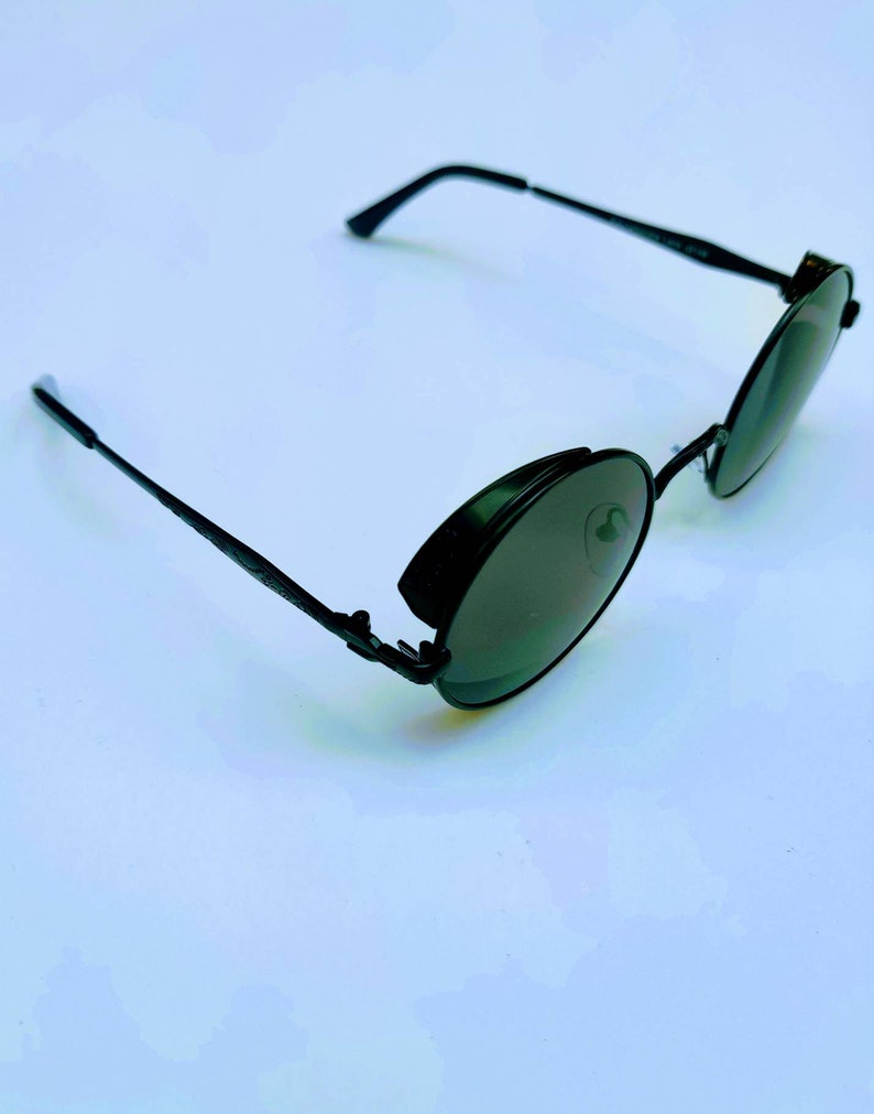 May include: Black round sunglasses with a metal frame and dark lenses. The sunglasses have a unique design with a hinge on the side of the frame.