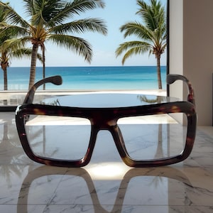 May include: Large, square-framed eyeglasses with a tortoiseshell pattern. The glasses are positioned on a reflective surface, with a tropical beach scene visible through the lenses. Palm trees and the ocean are in the background.