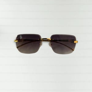 May include: Rimless sunglasses with a gold-colored frame and gradient lenses. The lenses transition from a dark brown at the top to a lighter shade at the bottom. The sunglasses have a rectangular shape and are set against a white background.