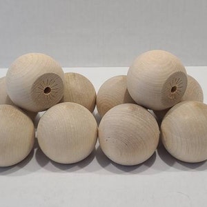 Puede incluir: Un conjunto de diez esferas de madera de color claro y superficie lisa dispuestas en grupo. Cada esfera presenta un pequeño orificio circular centrado con sutiles líneas grabadas que irradian alrededor de la abertura, mostrando la veta natural.