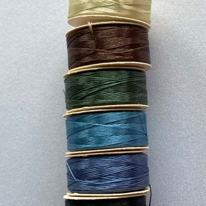 Puede incluir: Una pila de ocho carretes de hilo de varios colores, incluyendo crema, marrón, verde, azul, azul claro, negro y rojo.