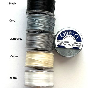 May include: Five spools of thread in black, grey, light grey, cream, and white. The spools are stacked on a wooden dowel. The label on the spool reads "One &bull; G Beads Thread 50yards TOHO CO.,LTD. 300denier Made in Japan."