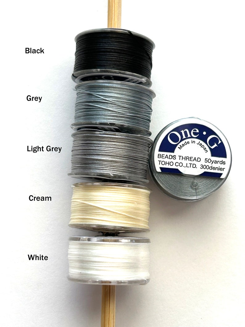 May include: Five spools of thread in black, grey, light grey, cream, and white. The spools are stacked on a wooden dowel. The label on the spool reads "One &bull; G Beads Thread 50yards TOHO CO.,LTD. 300denier Made in Japan."