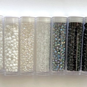 May include: Six clear plastic tubes filled with various types of seed beads. The beads are in different colors, including white, clear, iridescent, and black.
