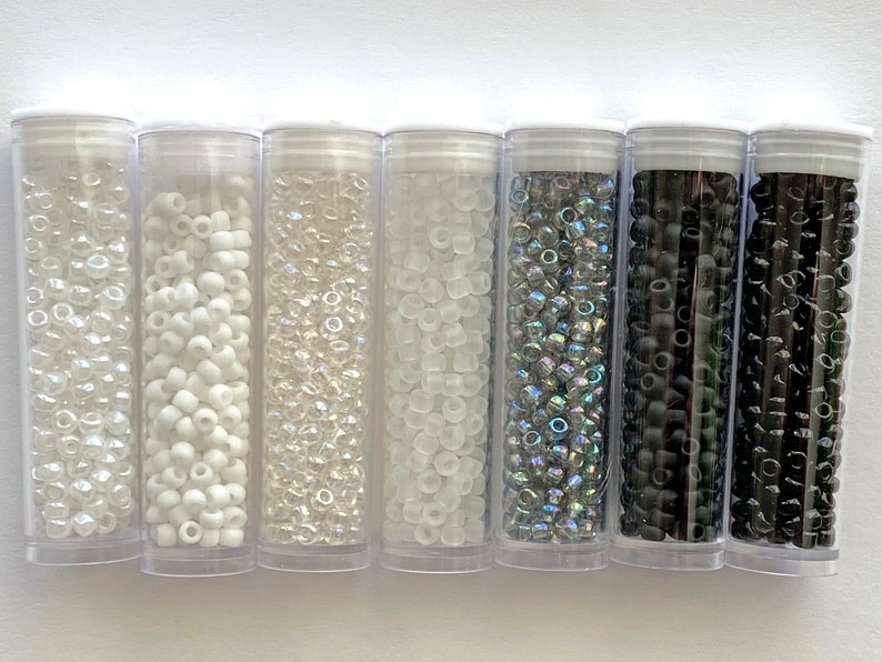 May include: Six clear plastic tubes filled with various types of seed beads. The beads are in different colors, including white, clear, iridescent, and black.
