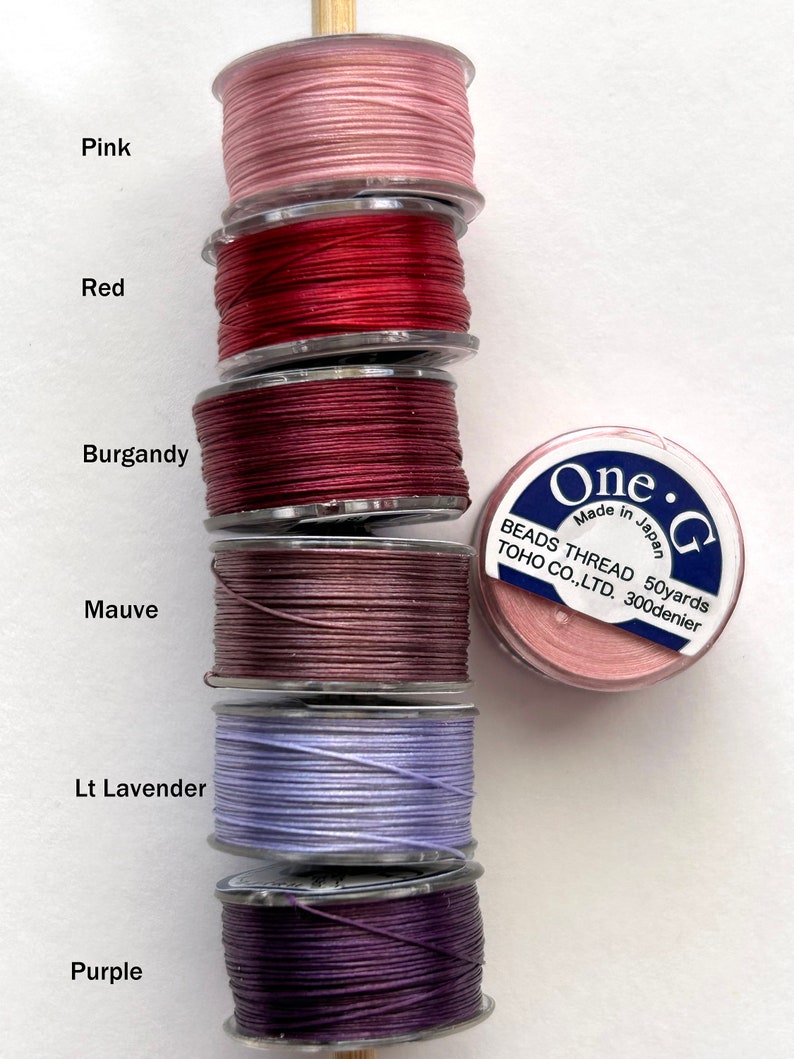 May include: Six spools of thread in various shades of pink, red, burgundy, mauve, light lavender, and purple. The spools are labeled with the color name and are stacked on top of each other. The spools are made by One-G Beads Thread TOHO CO.,LTD. and are 50 yards long and 300 denier.