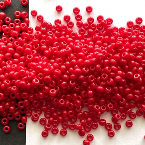 May include: A close-up of a collection of small, round, red beads. The beads are scattered on a white surface, with some spilling onto a dark surface. Each bead has a hole through the centre. The beads are a bright, solid red colour.