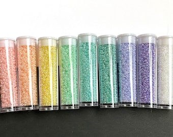 Może przedstawiać: Osiem przezroczystych plastikowych tub wypełnionych małymi, kolorowymi koralikami. Tuby ułożone są w rzędzie, prezentując spektrum pastelowych kolorów, w tym różowy, brzoskwiniowy, żółty, zielony, niebieski i fioletowy. Koraliki wydają się być koralikami nasiennymi, powszechnie używanymi do tworzenia biżuterii.