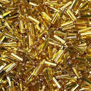 Può includere: Un primo piano mostra una fitta collezione di piccole perline cilindriche dorate. Le perline sono sparse, riflettendo la luce e creando un effetto scintillante. Questi componenti sono probabilmente utilizzati per la creazione di gioielli o altri progetti artigianali.