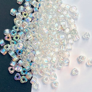 May include: A pile of clear, iridescent glass beads. The beads are a mix of square and triangular shapes, scattered on a white and black surface. The beads reflect a rainbow of colours, creating a shimmering effect. Suitable for jewellery making and crafts.