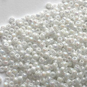 Peut inclure: Un grand tas de perles de rocaille blanches iris&eacute;es. Les perles sont petites et rondes, avec une forme l&eacute;g&egrave;rement irr&eacute;guli&egrave;re. Elles ont un &eacute;clat nacr&eacute; et sont parfaites pour la fabrication de bijoux.