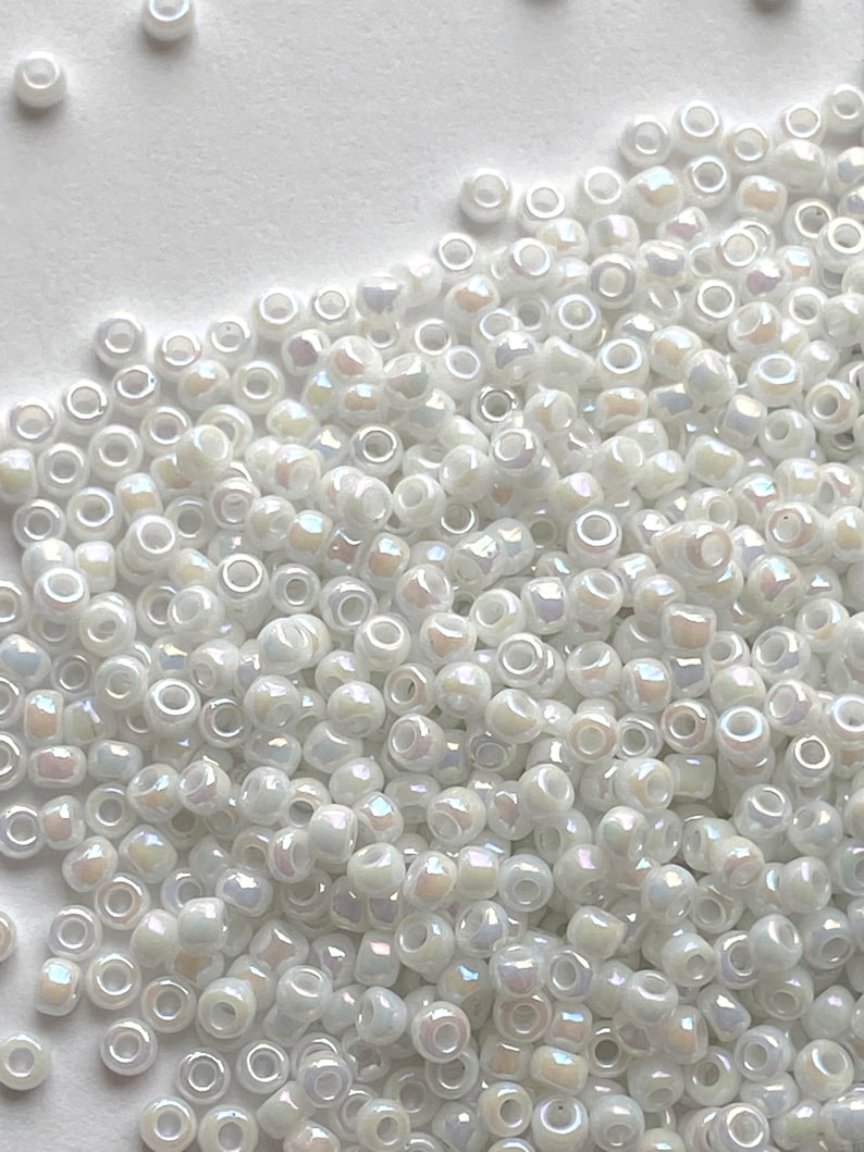 Peut inclure: Un grand tas de perles de rocaille blanches iris&eacute;es. Les perles sont petites et rondes, avec une forme l&eacute;g&egrave;rement irr&eacute;guli&egrave;re. Elles ont un &eacute;clat nacr&eacute; et sont parfaites pour la fabrication de bijoux.
