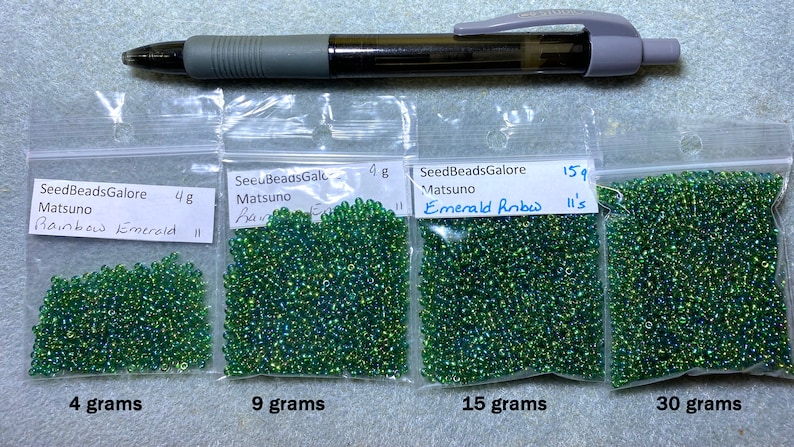 Peut inclure: Quatre sachets de perles de rocaille vertes &eacute;tiquet&eacute;s avec le nom de la marque "SeedBeadsGalore" et le poids des perles en grammes. Les sachets sont &eacute;tiquet&eacute;s avec la couleur des perles "Rainbow Emerald" et "Emerald Rnbw".