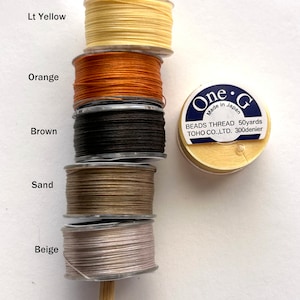 May include: Spools of thread in various colors, including light yellow, orange, brown, sand, and beige. The spools are stacked on a wooden dowel. A label on a separate spool reads "One G Made in Japan BEADS THREAD 50yards TOHO CO.,LTD. 300denier".