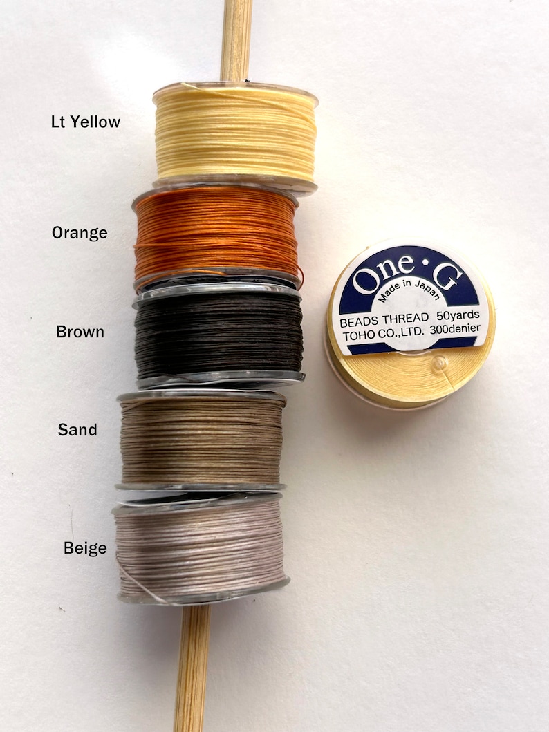 May include: Spools of thread in various colors, including light yellow, orange, brown, sand, and beige. The spools are stacked on a wooden dowel. A label on a separate spool reads "One G Made in Japan BEADS THREAD 50yards TOHO CO.,LTD. 300denier".