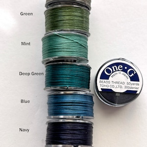 May include: Six spools of thread in various shades of green, blue, and black. The spools are labeled with the colors: LT Khaki, Green, Mint, Deep Green, Blue, and Navy. The spools are arranged vertically on a wooden dowel. A label on the right side of the image reads "One-G Beads Thread 50yards TOHO CO.,LTD. 300denier Made in Japan."