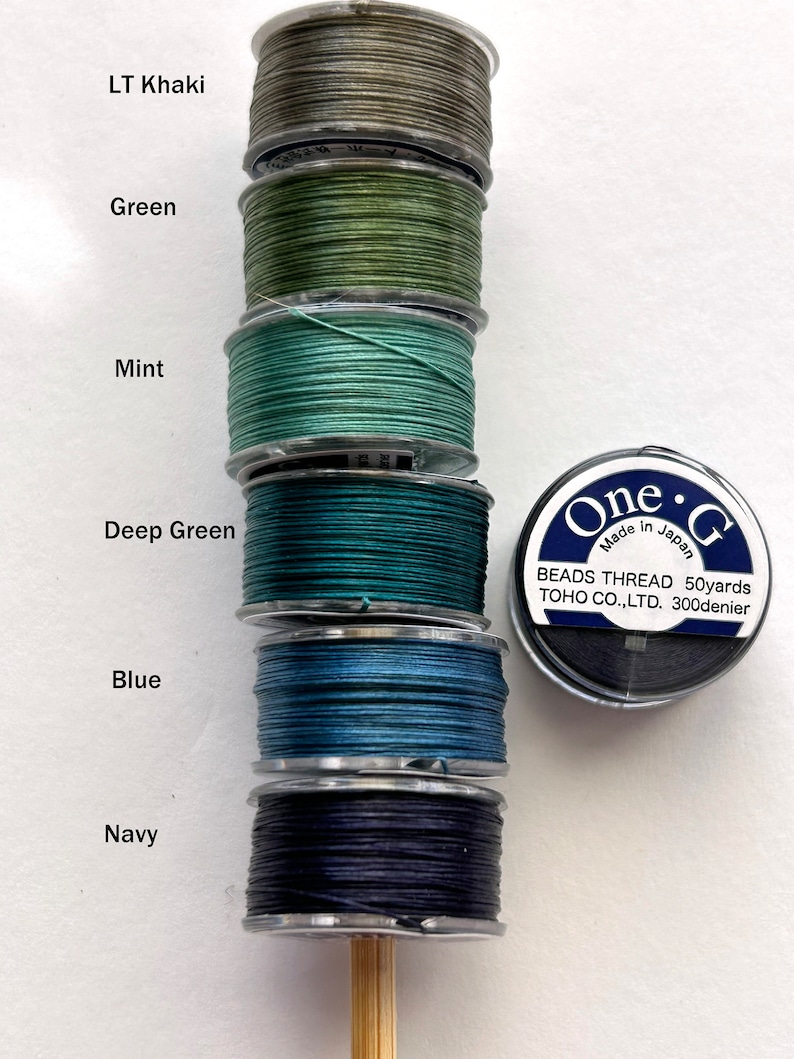 May include: Six spools of thread in various shades of green, blue, and black. The spools are labeled with the colors: LT Khaki, Green, Mint, Deep Green, Blue, and Navy. The spools are arranged vertically on a wooden dowel. A label on the right side of the image reads "One-G Beads Thread 50yards TOHO CO.,LTD. 300denier Made in Japan."