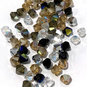 May include: A collection of faceted bicone beads in a mix of clear, black, and various shades of brown and gold. Each bead features a multi-faceted surface designed to reflect light, with some showing iridescent or metallic finishes throughout.