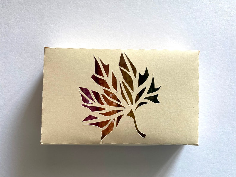 May include: A beige box with a cut-out design of a maple leaf in shades of pink, orange, brown, and green.