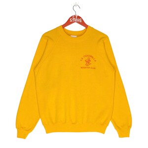 May include: Yellow sweatshirt with a red and orange graphic of a person riding a horse with the text "La Cuadrilla Booster Club".
