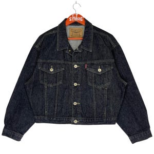 May include: A dark blue denim jacket with a button closure and two front pockets. The jacket has a Big John label on the left chest.