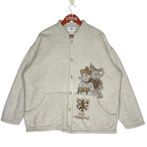 May include: A light gray button-up sweatshirt with a cartoon illustration of two animals wearing winter hats and scarves. The sweatshirt has a large pocket on the front with the text "The Champions" embroidered on it.