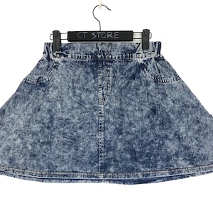 May include: A blue denim skirt with a white acid wash pattern. The skirt has a high waist and a flared silhouette. The skirt has a small logo patch on the right side.