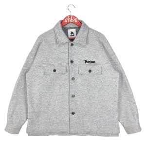 May include: A gray long-sleeved button-up shirt with a black embroidered logo that says "MOZ" and a small moose graphic. The shirt has two flap pockets on the chest.