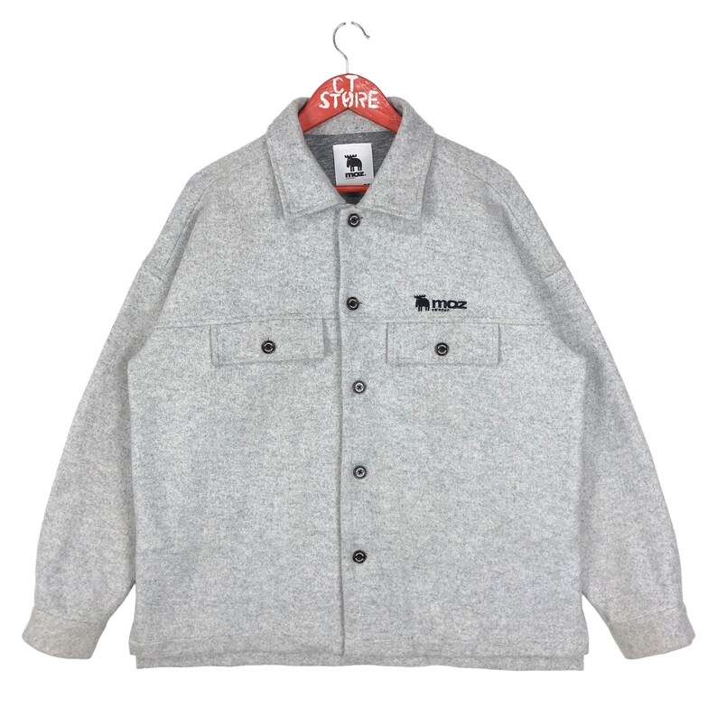 May include: A gray long-sleeved button-up shirt with a black embroidered logo that says "MOZ" and a small moose graphic. The shirt has two flap pockets on the chest.