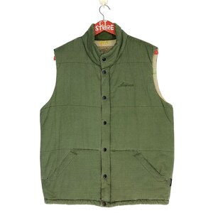 May include: A green quilted vest with a button-up front and a small pocket on the left side. The vest has a high collar and is made of a lightweight fabric. The vest has the word "Alvar" embroidered on the front.