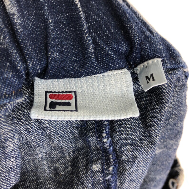 Op de afbeelding: Een wit label met een rood en blauw logo met de tekst "FILA" erop. Het label is bevestigd aan een blauw denim kledingstuk. De maat "M" staat op een wit label dat aan het kledingstuk is bevestigd.