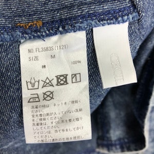 Op de afbeelding: Een wit waslabel met wasinstructies voor een denim kledingstuk. Het label heeft de tekst "NO. FL3583S (1121)", "SIZE M", "綿 100%" en een reeks symbolen die aangeven hoe het kledingstuk gewassen, gebleekt, gedroogd en gestreken moet worden.