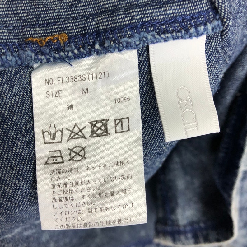 Op de afbeelding: Een wit waslabel met wasinstructies voor een denim kledingstuk. Het label heeft de tekst "NO. FL3583S (1121)", "SIZE M", "綿 100%" en een reeks symbolen die aangeven hoe het kledingstuk gewassen, gebleekt, gedroogd en gestreken moet worden.