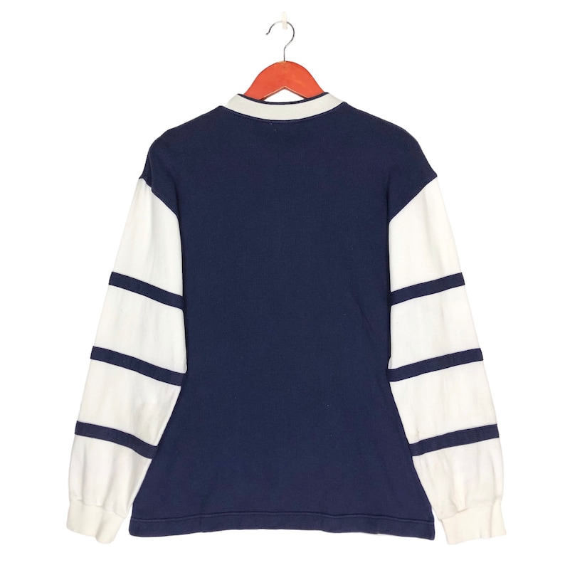 May include: A navy blue long-sleeved shirt with white striped sleeves. The shirt has a collared neckline and a button closure.